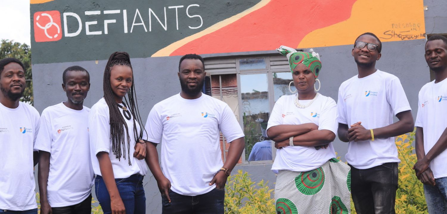 Unidos-team-Nakivale-refugee-settlement-Uganda-regenerativ-empowerment Unidos team, Nakivale Refugee Settlement, Uganda, seven people standing in front of building with lettering DEFIANTS